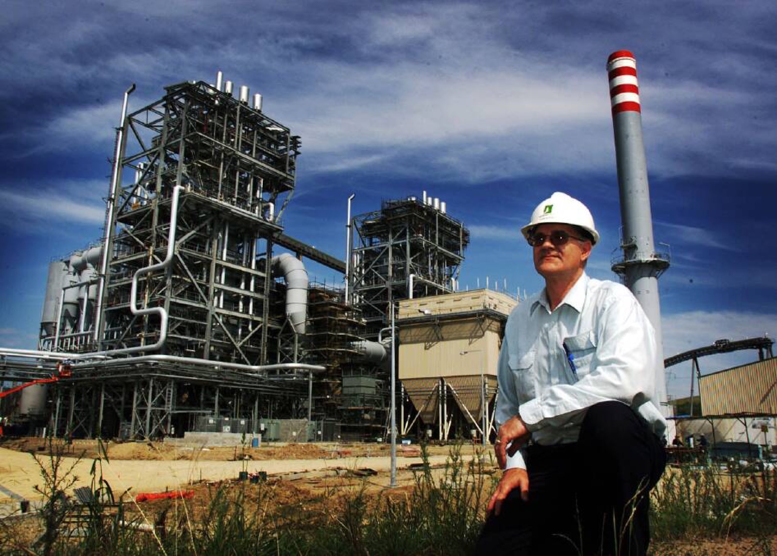 Sam in a white shirt and hard hat, kneeling on rough ground in front of the Redbank power station, its boiler structures and red-and-white striped chimney rising behind him under a deep blue sky.