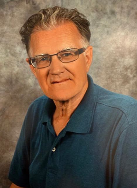 Studio portrait of Sam in his later years: silver-grey hair, glasses, and a teal polo shirt, with a soft smile.